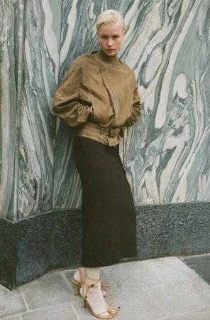 Adult woman in tan jacket and dark skirt poses against marble patterned wall wearing strappy heels with ankle ties in a fashion shot.