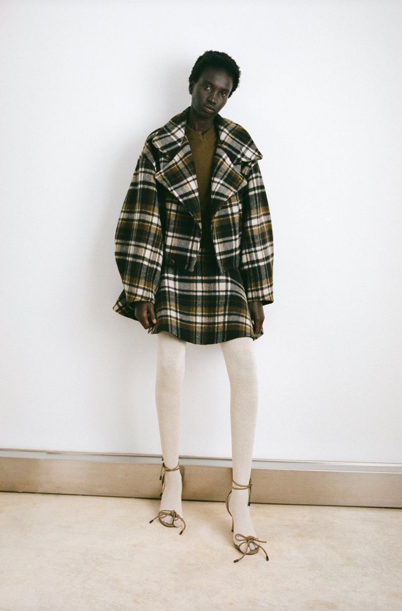 Fashion model poses in a green and brown plaid mini skirt and jacket, accessorized with beige tights and strappy heels against a white wall.