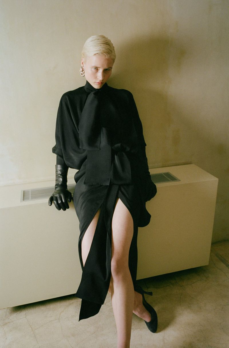 Indoor portrait of a blonde adult woman wearing a black dress and black gloves, posed against an off-white wall and radiator.