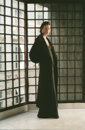 Model wearing a long black coat standing in front of a grid window for Petar Petrov A/W 25 collection.