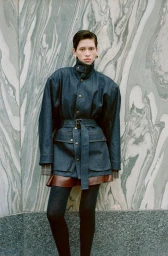 Model standing against patterned marble wall wearing a belted dark jacket and skirt