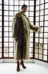 Model wearing a long textured faux fur coat standing against a large grid window panel