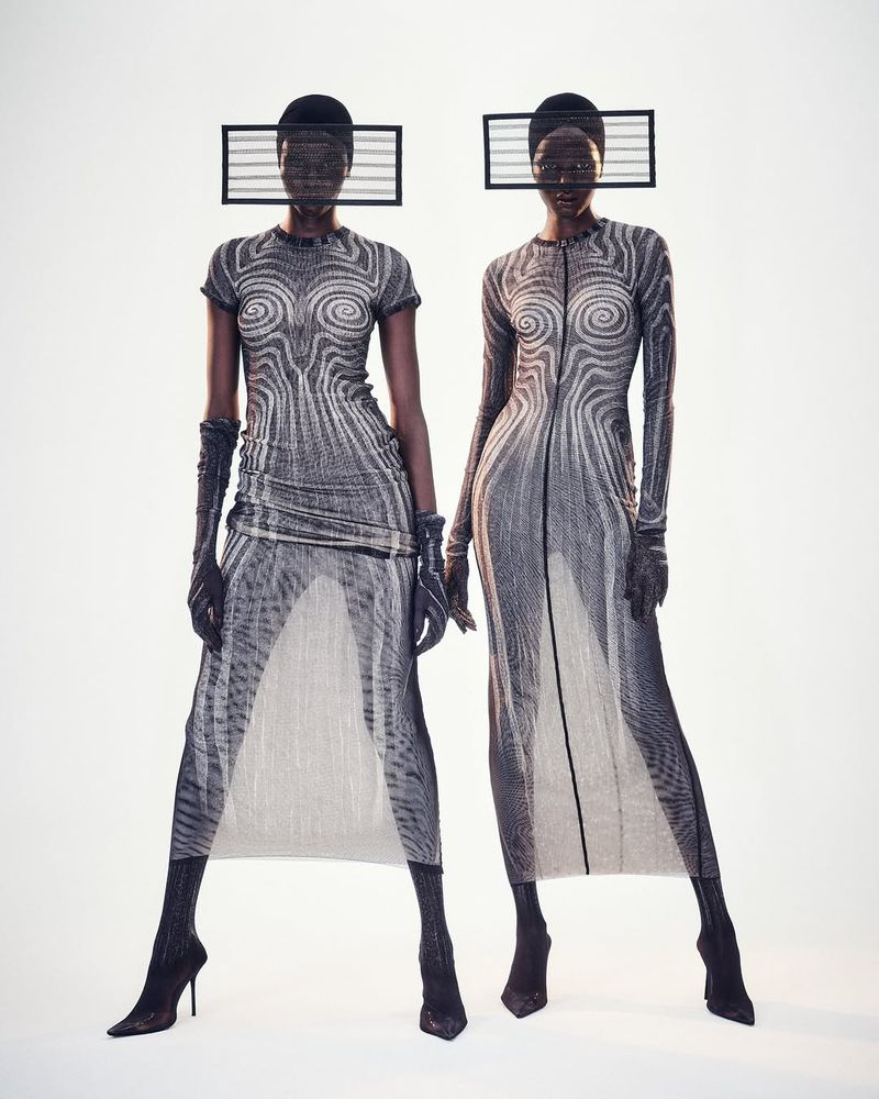 Two Black female fashion models pose in gray-patterned gowns with geometric headwear against a bright background in a studio shot.