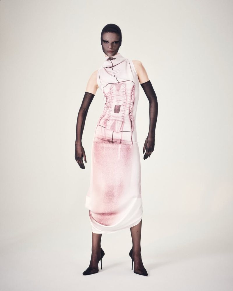 Fashion shot of a slender adult female wearing a high neck, sleeveless pink and white dress with black heels and arm gloves in a studio.