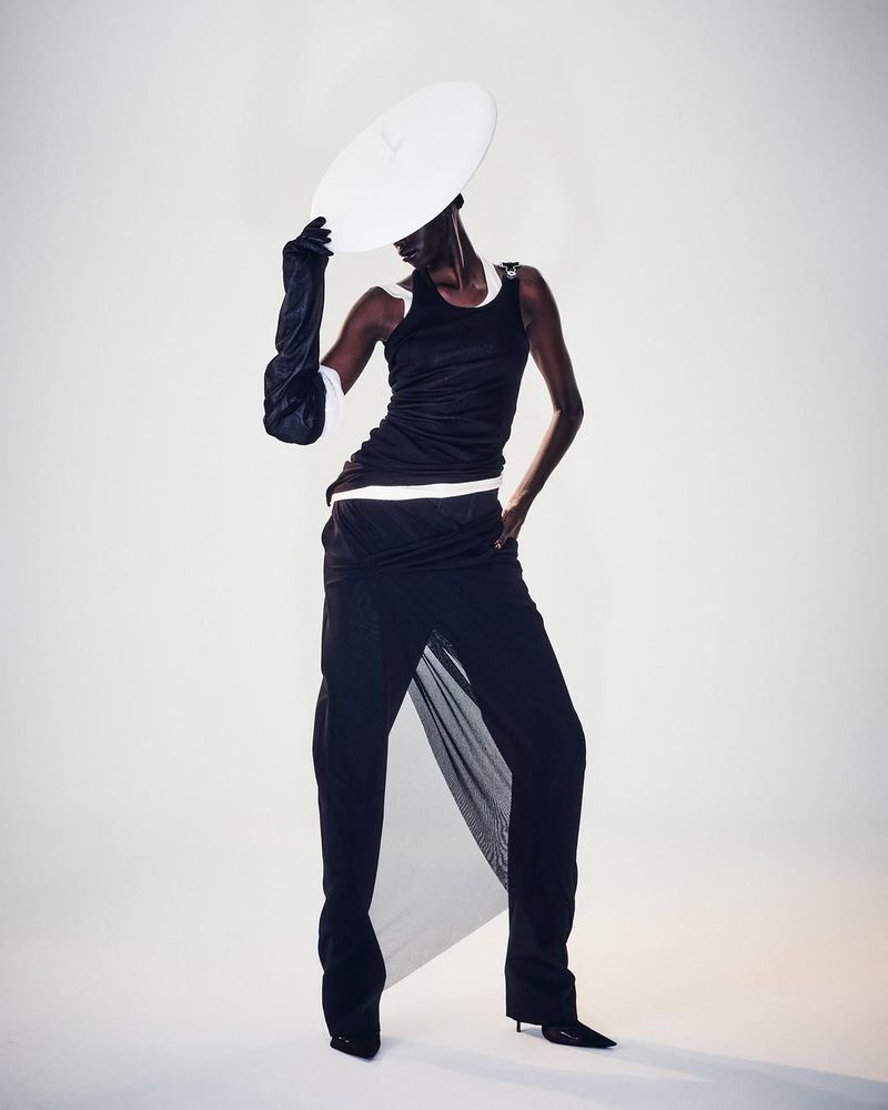 A woman poses in a black tank top, flowing black pants, opera gloves, and a white disc hat against a solid white backdrop.