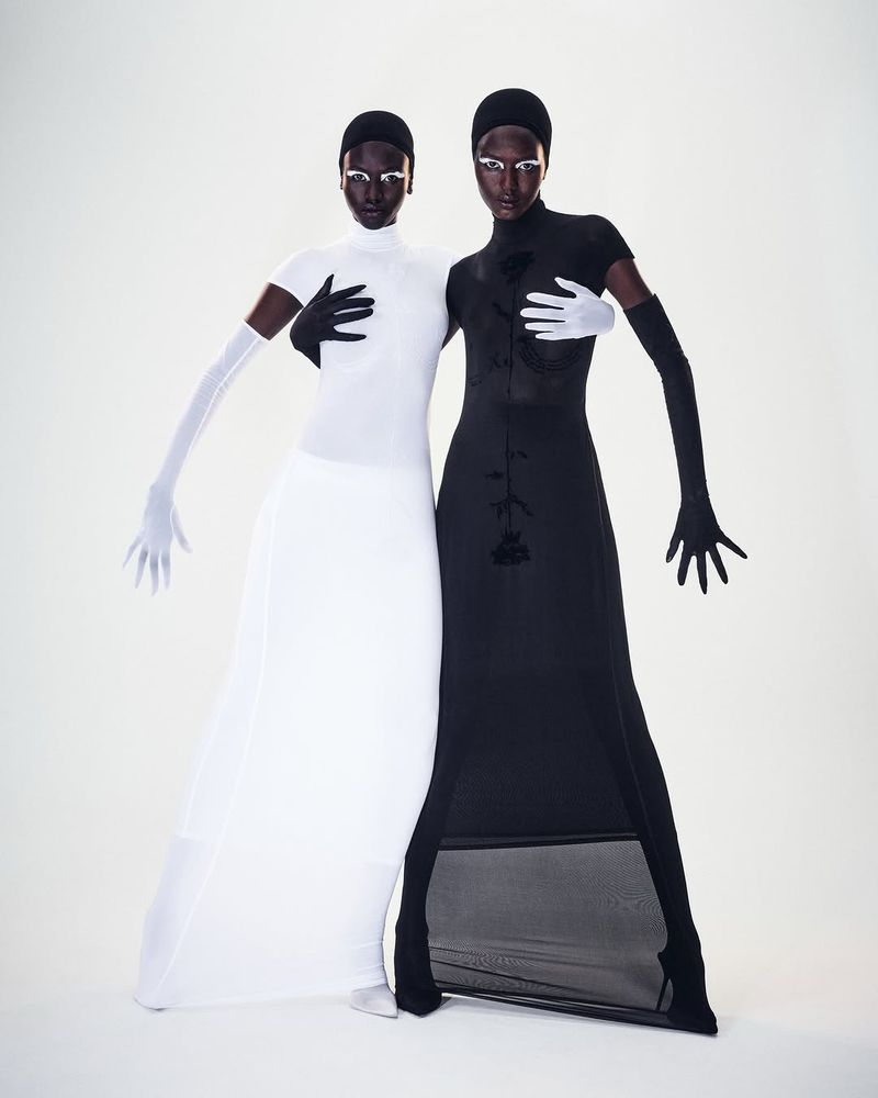 Two fashion models pose in contrasting black and white gowns and matching gloves and headwear in a studio setting.