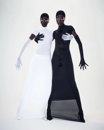 Two fashion models pose in contrasting black and white gowns and matching gloves and headwear in a studio setting.