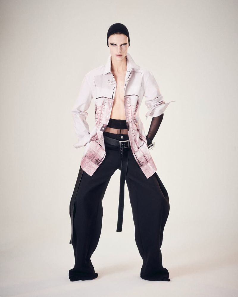 A fashion model in a long-sleeved shirt, wide-leg pants, and head covering poses against a light background in a full-length studio shot.
