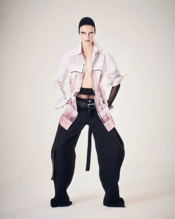 A fashion model in a long-sleeved shirt, wide-leg pants, and head covering poses against a light background in a full-length studio shot.