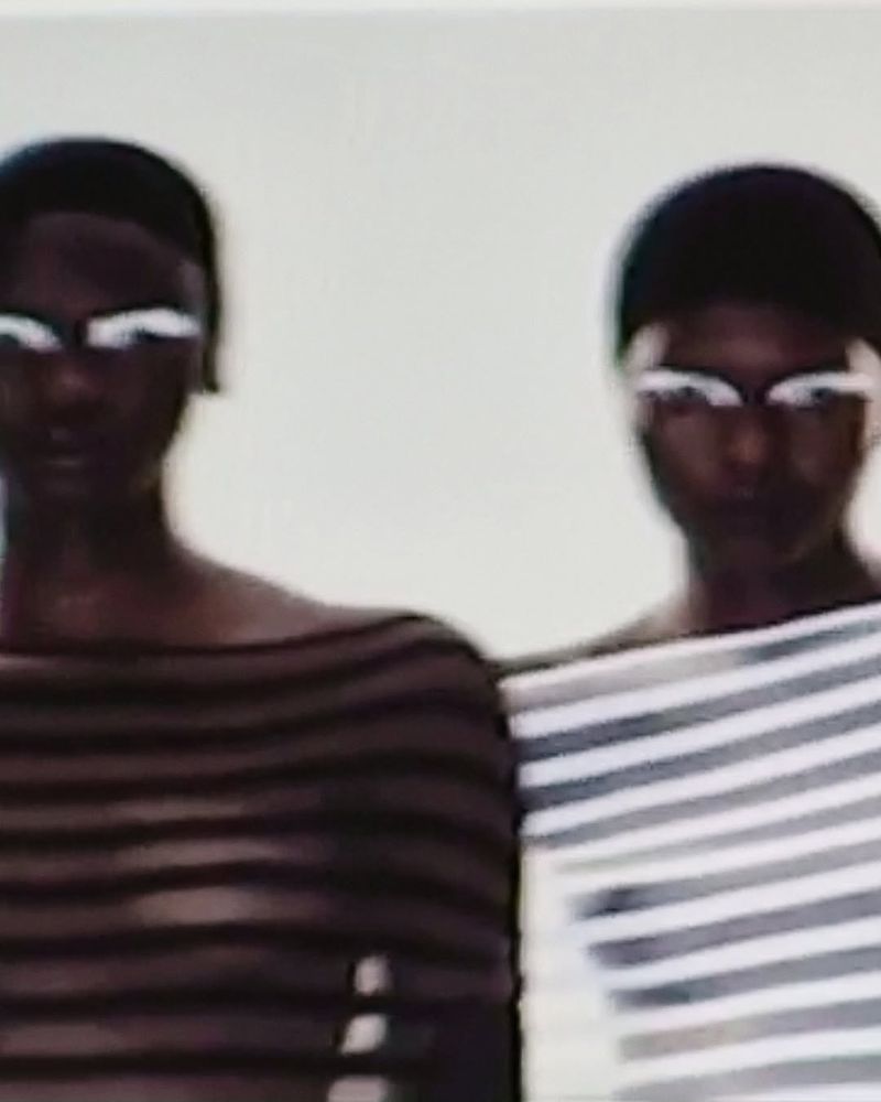 Two adult women with dark skin and white eye makeup in off-shoulder striped tops against a plain background in a portrait orientation.