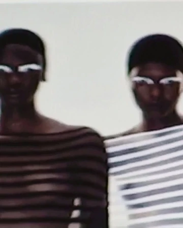 Two adult women with dark skin and white eye makeup in off-shoulder striped tops against a plain background in a portrait orientation.