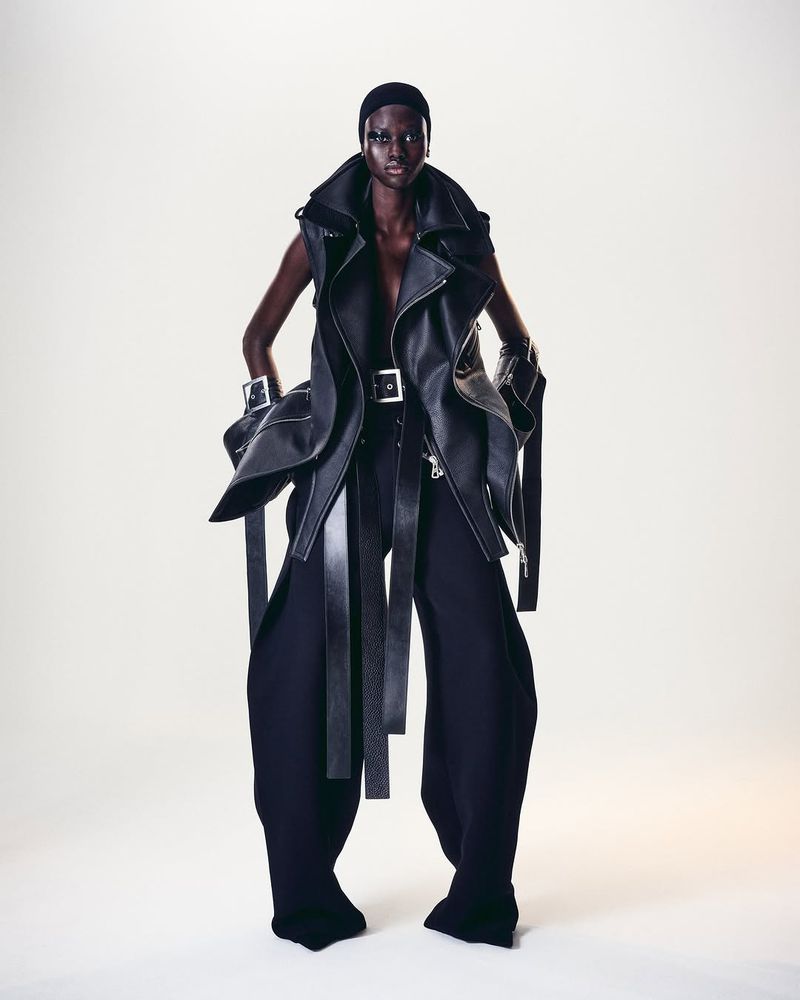 A striking fashion portrait features a Black model in an avant-garde black leather vest, wide black pants, and bold silver cuff bracelets.