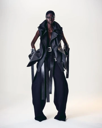 A striking fashion portrait features a Black model in an avant-garde black leather vest, wide black pants, and bold silver cuff bracelets.