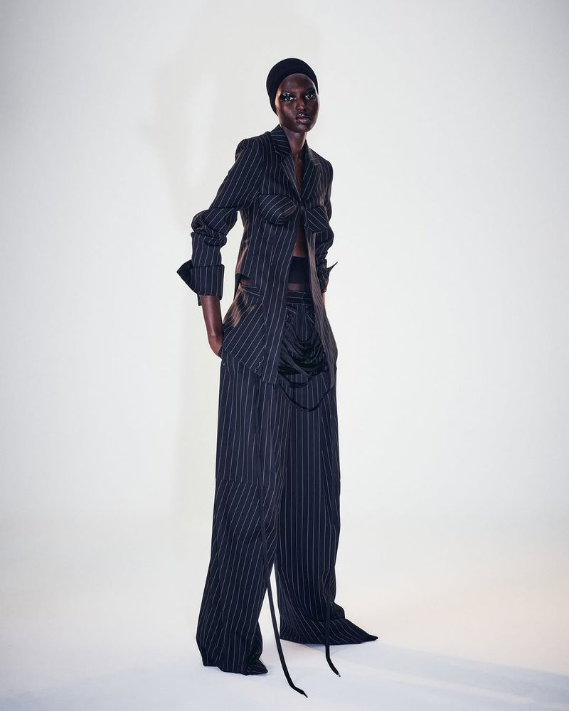 A Black fashion model strikes a studio pose wearing a black and white pinstripe blazer and trousers with a black head covering.