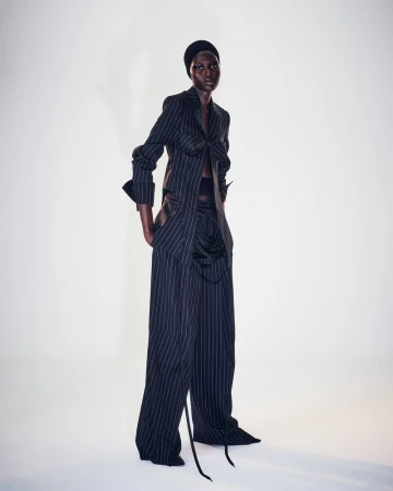 A Black fashion model strikes a studio pose wearing a black and white pinstripe blazer and trousers with a black head covering.