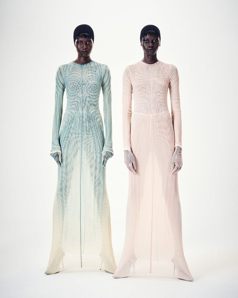 Two dark-skinned women in long, form-fitting dresses with intricate patterns stand side-by-side against a bright, neutral backdrop in a fashion shot.