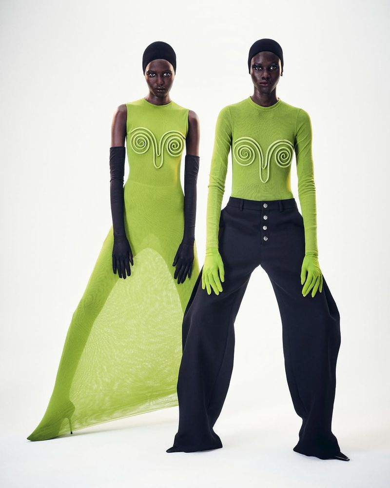 Two dark-skinned fashion models pose in a photo studio. One model wears a green dress with black gloves and the other wears green top and black pants.