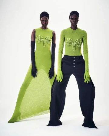 Two dark-skinned fashion models pose in a photo studio. One model wears a green dress with black gloves and the other wears green top and black pants.