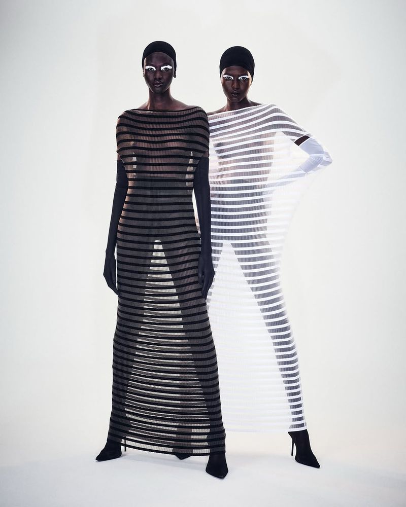 Two women pose in horizontal-striped sheer dresses against a white backdrop, accentuating a striking visual display of fashion and style.