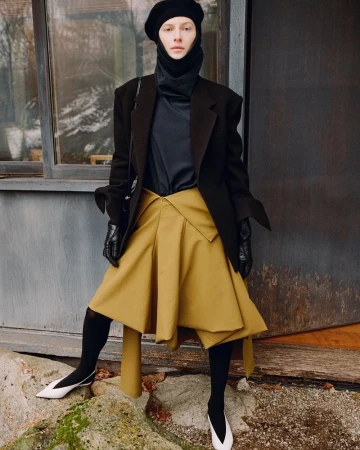 Fashionable person posing in a black jacket, tan skirt, and beret, standing on rocks in front of a gray building. Outdoor fashion.