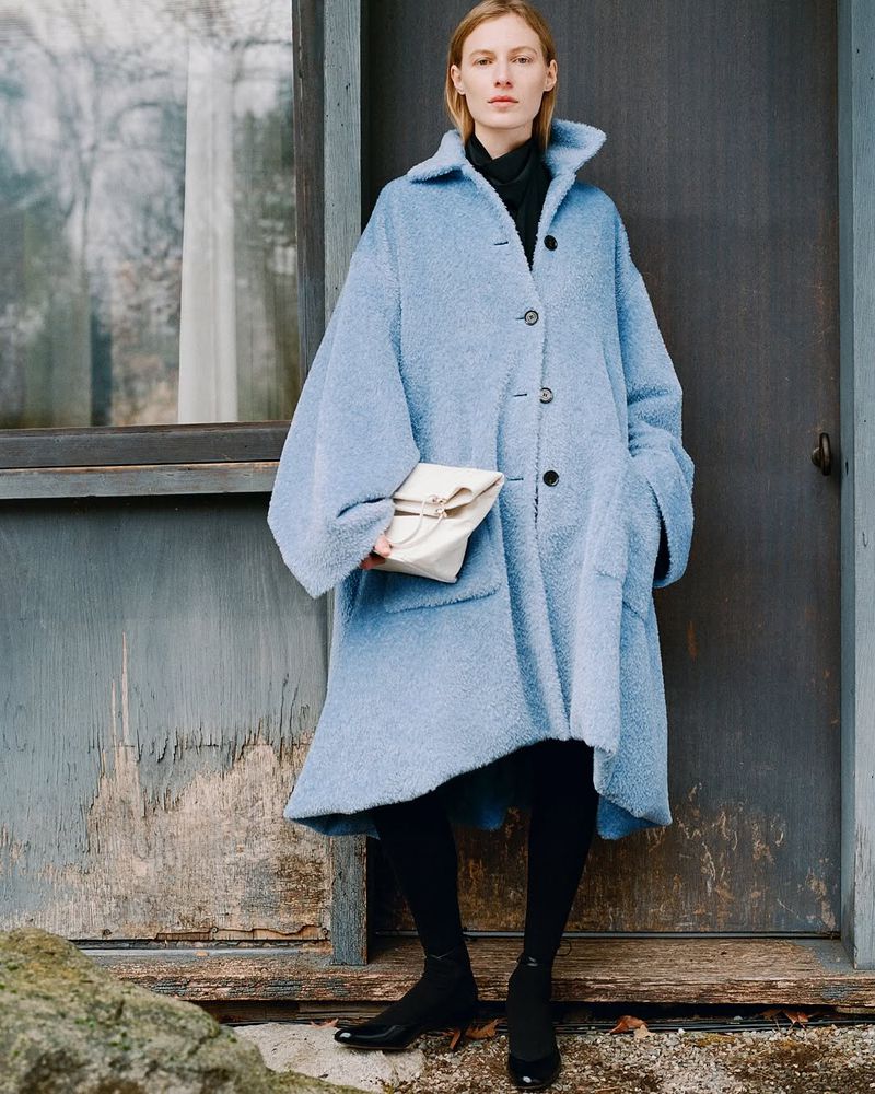 Model wearing a loose blue textured coat holding a white clutch standing in front of a rustic door and window frame