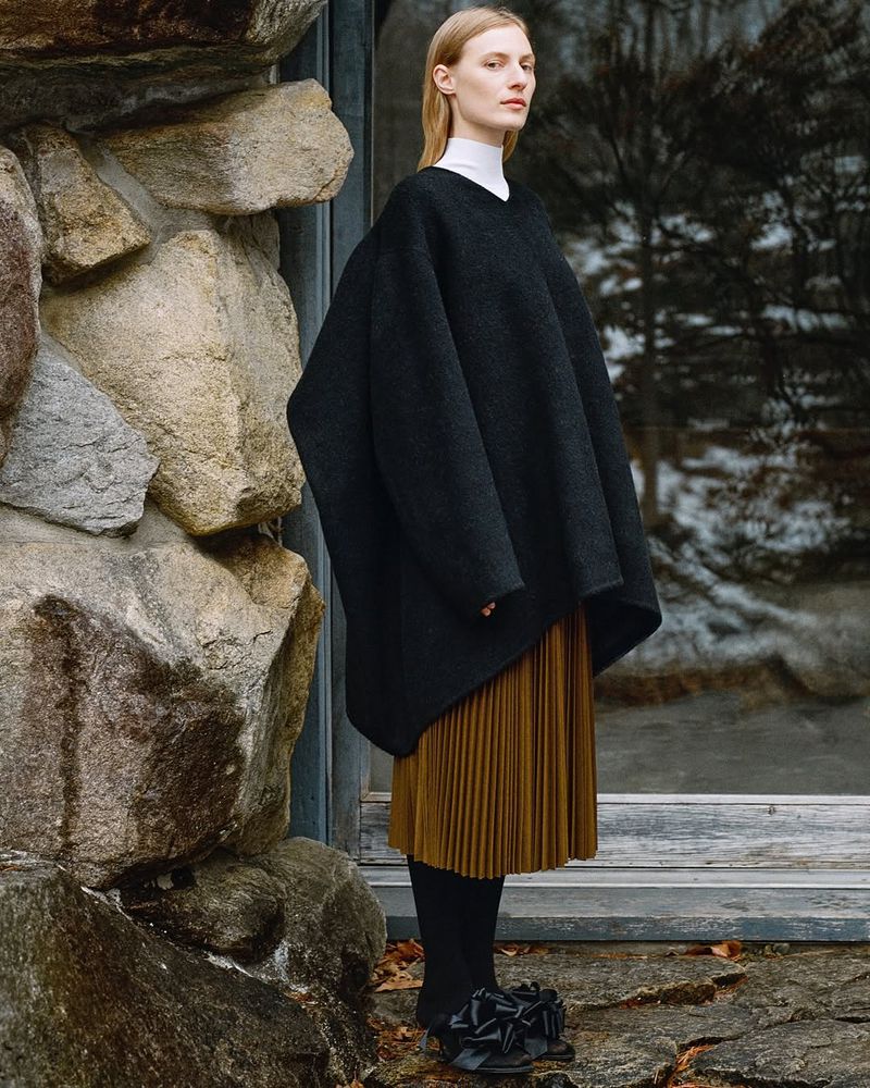 A fair-skinned woman models a black poncho, pleated skirt, and sculptural shoes in front of a rock wall and reflective glass windows.