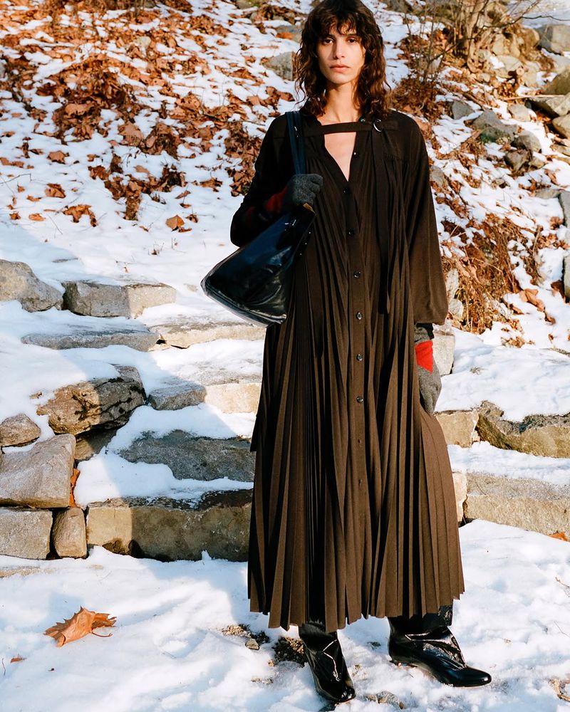 A woman with curly hair stands on snow-covered steps outside, wearing a long, brown, pleated dress, black boots, and carrying a black bag.