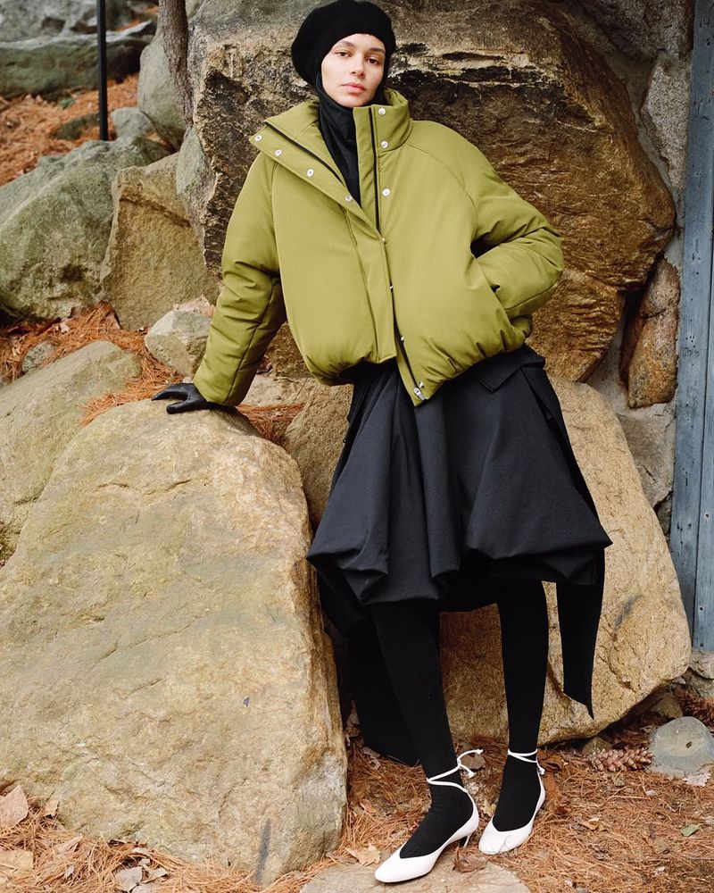 A woman in a puffy olive jacket, black skirt, tights, and beret leans against a large rock formation in an outdoor setting.