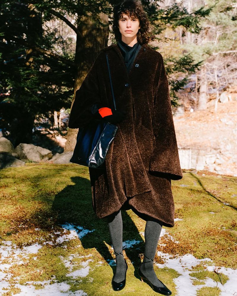 A woman in a brown coat stands in a forest clearing, modeling fashion with natural lighting and a blurred background featuring trees and foliage.