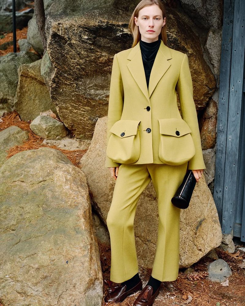 A model poses outdoors in a tailored yellow suit with oversized pocket details, paired with leather loafers and a black bag.
