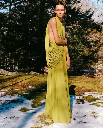 Model standing outdoors in a flowing olive pleated dress against a snowy ground and dark trees for Proenza Schouler