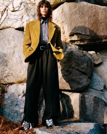 A model poses against a rock wall wearing a mustard-colored corduroy blazer, black wide-leg pants, and silver bow-embellished shoes.