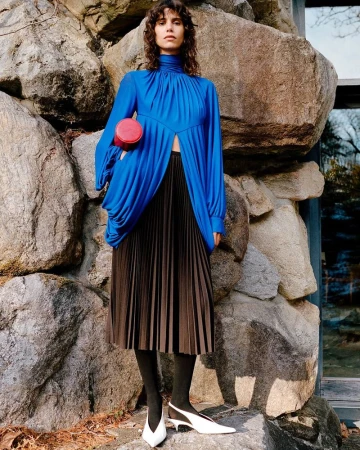 Model standing outdoors against large rock formations wearing a royal blue draped top and dark pleated skirt.