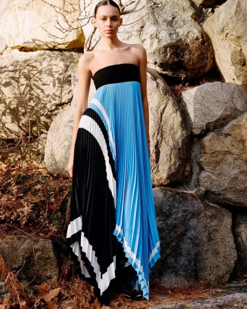 Model standing against large rocky boulders wearing a strapless pleated blue, black, and white dress for Proenza Schouler.