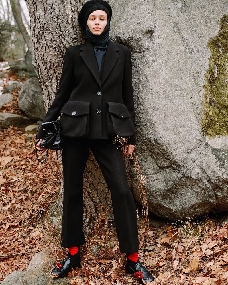 A stylish young woman in a black blazer, pants, and beret poses outdoors next to a large rock, creating a moody fashion image.