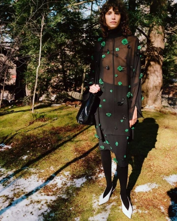 A slender adult female stands in a grassy, wooded landscape wearing a long-sleeved black semi-sheer floral-embellished dress and white heels.