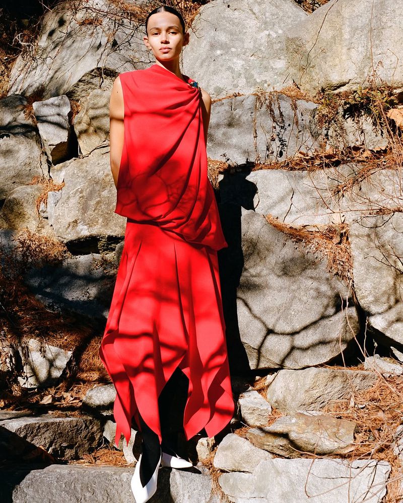 Adult woman in a flowing red dress and white shoes poses in front of a stone wall outdoors in natural light, fashion editorial.