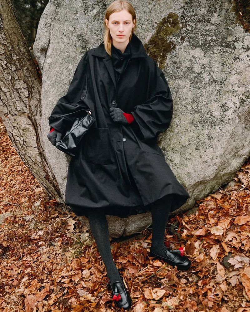 A fair-skinned woman in a black coat poses next to a large rock outdoors surrounded by fallen leaves in a fashion editorial shot.