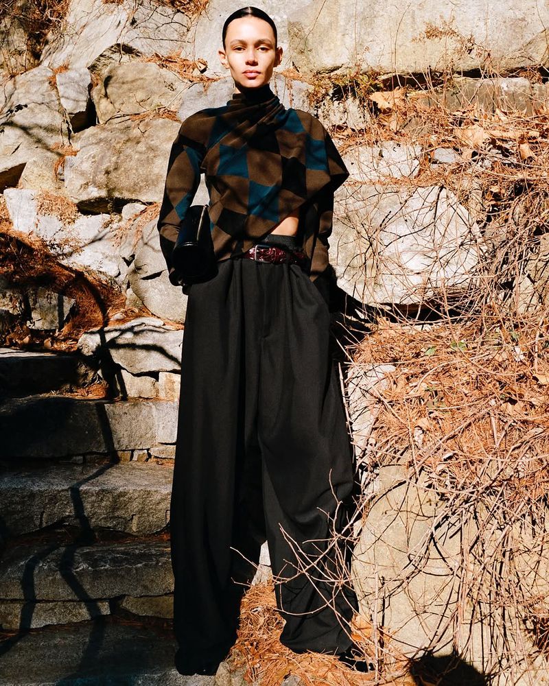 A woman poses in a patterned top and black pants against a stone wall and branches in an outdoor setting on a sunny day.