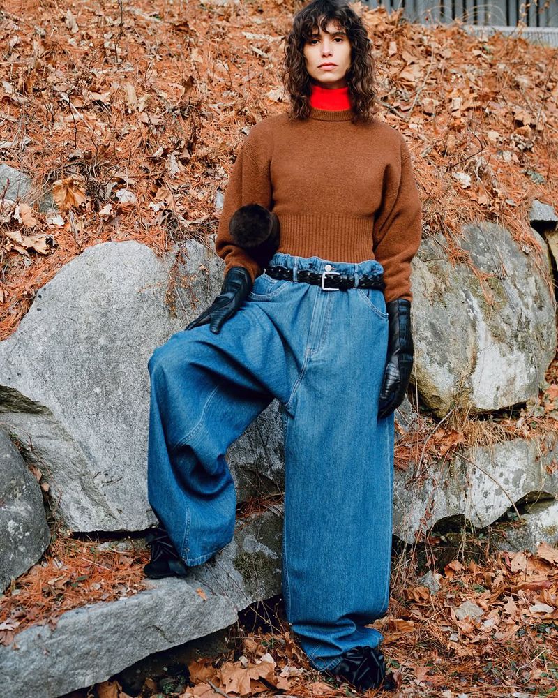 Fashionable woman with curly hair wearing a brown sweater and baggy blue jeans, posing against a rock wall outdoors in a natural setting.