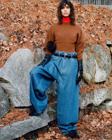 Fashionable woman with curly hair wearing a brown sweater and baggy blue jeans, posing against a rock wall outdoors in a natural setting.
