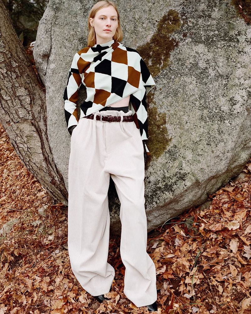 Adult female fashion model poses in a brown, white, and black patterned top and white corduroy pants beside a large rock in nature.
