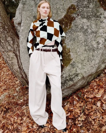 Adult female fashion model poses in a brown, white, and black patterned top and white corduroy pants beside a large rock in nature.
