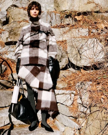 Woman in a brown and white plaid jacket and skirt stands in an outdoor setting with stone steps and brown vines.