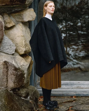 Model standing outdoors wearing an oversized black textured top and a pleated brown skirt near a stone wall.