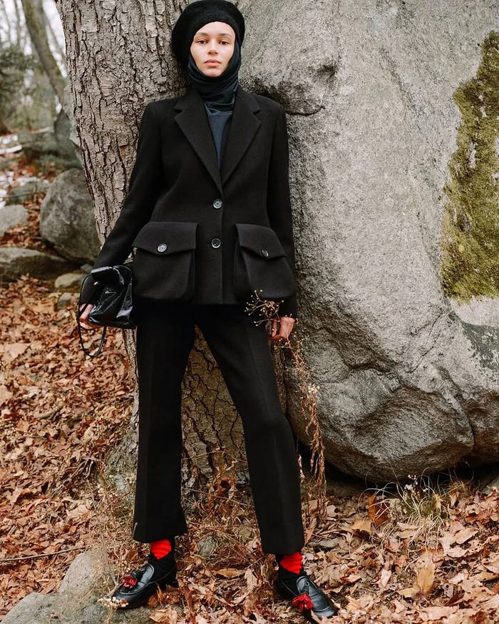 Model wearing a black tailored jacket, trousers, and head covering leaning against a large rock in an outdoor woodland setting.