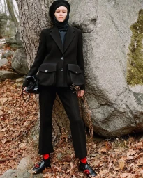 Model wearing a black tailored jacket, trousers, and head covering leaning against a large rock in an outdoor woodland setting.