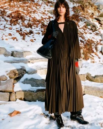 Model standing outdoors on a snowy rocky path wearing a dark pleated long dress and carrying a black leather handbag.