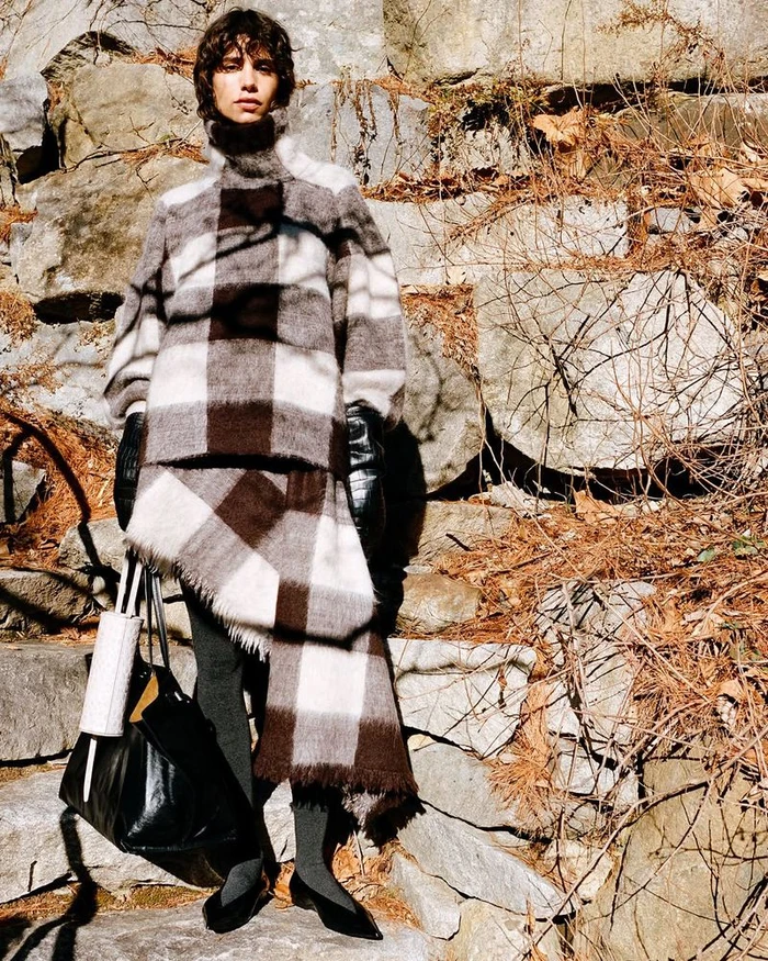 Model wearing a brown and white plaid turtleneck standing against a rocky outdoor background