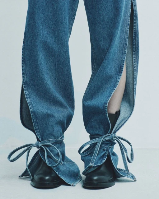 Close-up of denim pants with split hems tied around black leather shoes, showcasing a modern fashion design against a plain background.